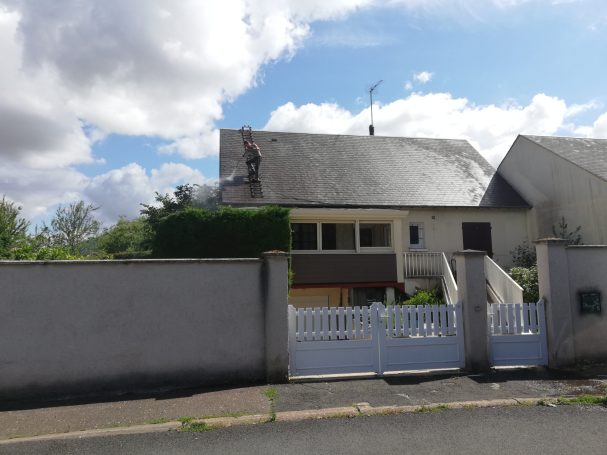 Maison à un étage avec un toit en pente, entourée de verdure, sous un ciel nuageux.